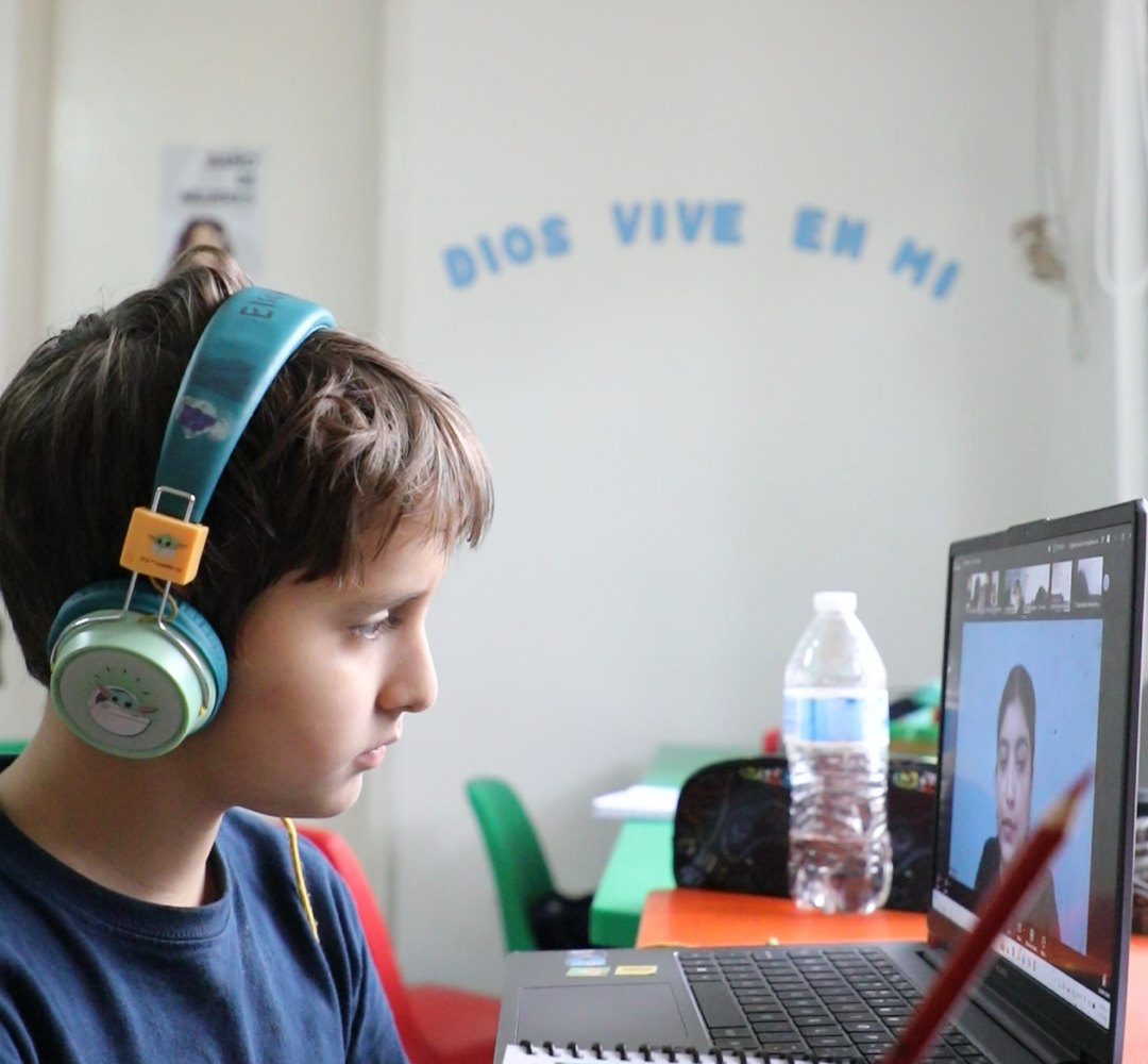 Un alumno de primaria del Instituto Zoe sigue su clase en línea con sus audífonos, bajo el letrero «Dios vive en mí»