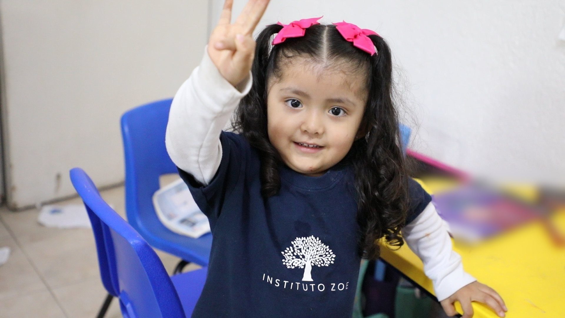 Una alumna de kínder del Instituto Zoe sonríe y levanta tres dedos desde su salón