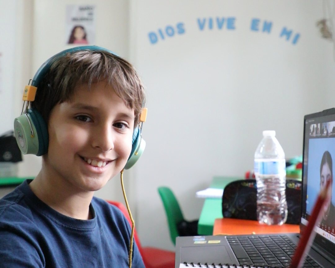 Un alumno de primaria del Instituto Zoe sonríe junto a su computadora; en la pared del salón se lee «Dios vive en mí»