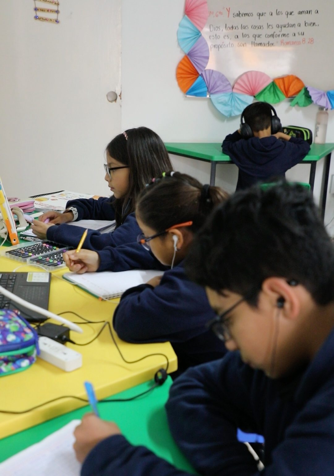 Alumnos de primaria del Instituto Zoe trabajan en sus cuadernos; en la pared, el versículo de Romanos 8:28