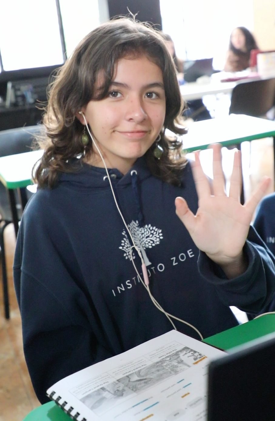 Una alumna de secundaria del Instituto Zoe sonríe y saluda con la mano desde su lugar, con su cuaderno de inglés abierto